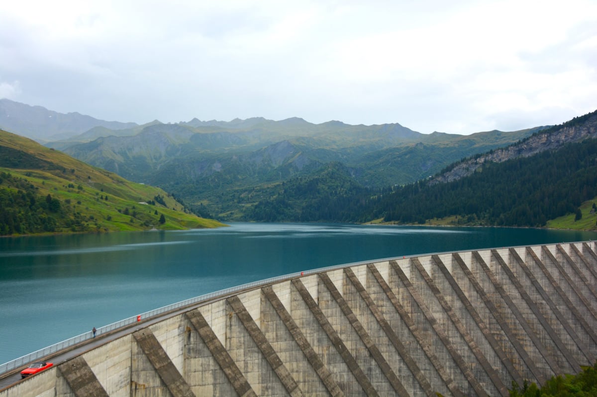 Large concrete dam spanning across a turquoise alpine lake surrounded by forested mountains and green hillsides.