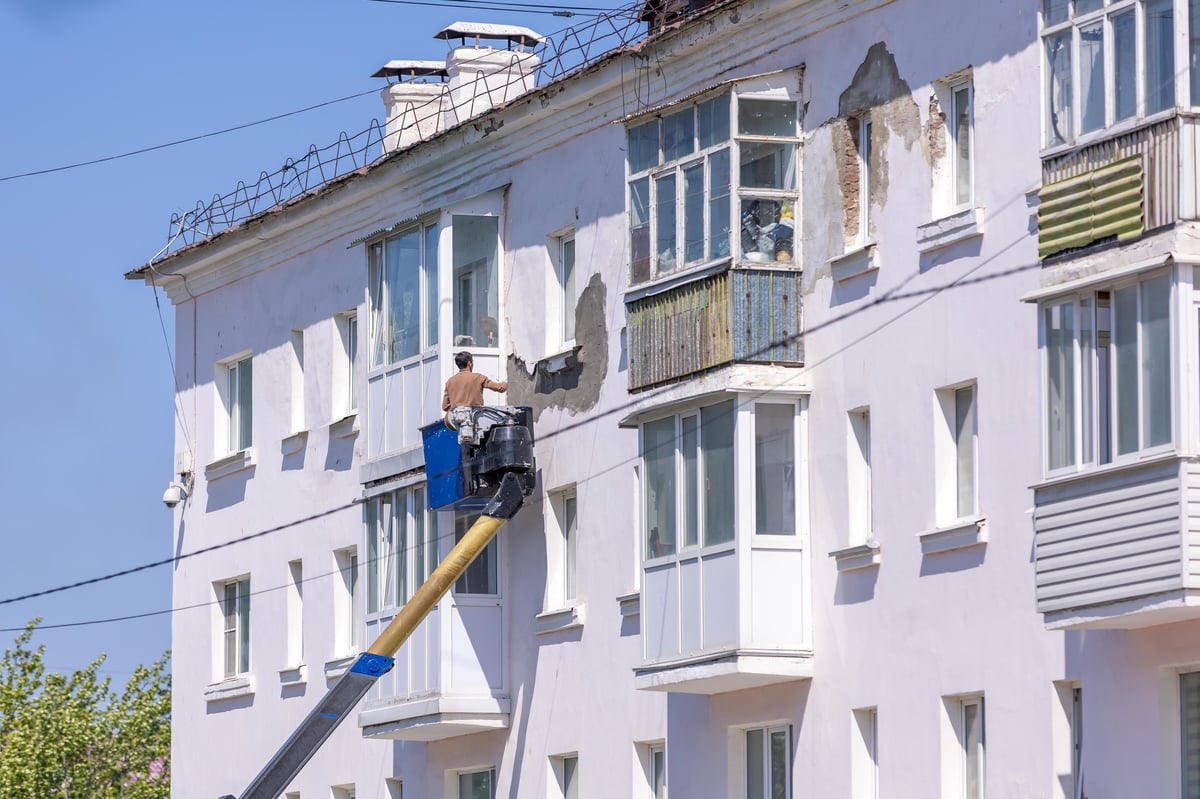 Worker on elevated cherry picker platform maintaining exterior of white apartment building facade
