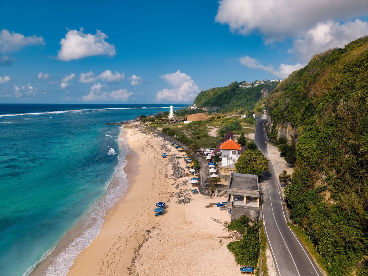 Aerial view of a coastal road along a sandy beach with turquoise ocean, cliffs covered in green vegetation, and buildings including a lighthouse on the shoreline under blue sky with white clouds.