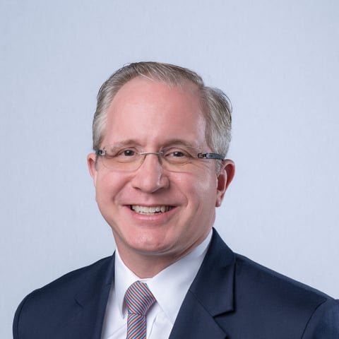 Professional headshot of a smiling middle-aged man with gray hair, glasses, wearing a dark suit and striped tie against a light blue background