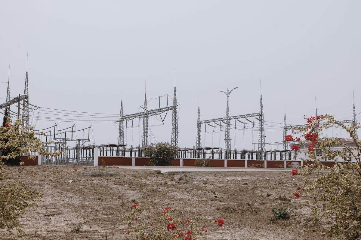 Industrial high-voltage electrical substation with steel transmission towers, power lines, and concrete structures on barren terrain under cloudy sky.