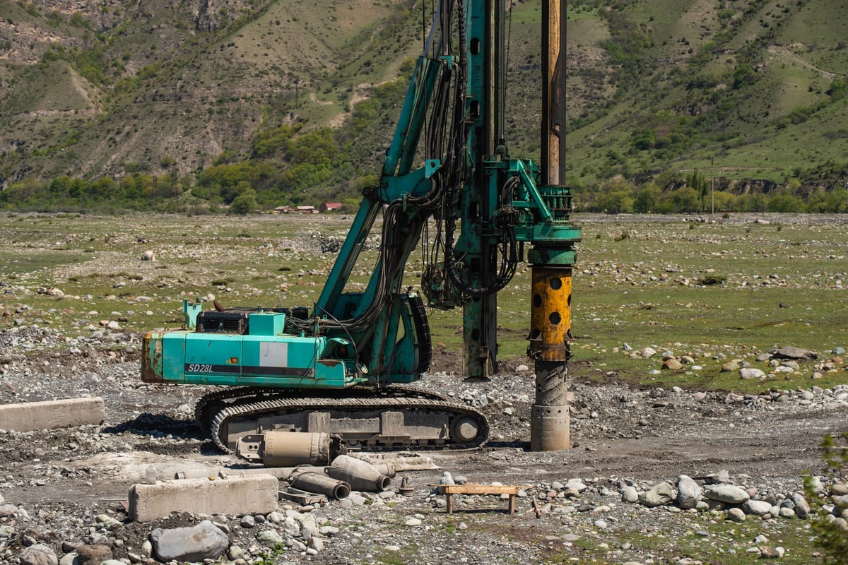 Turquoise and yellow drill rig with extended drilling arm operating in a mountain valley landscape with rocky terrain