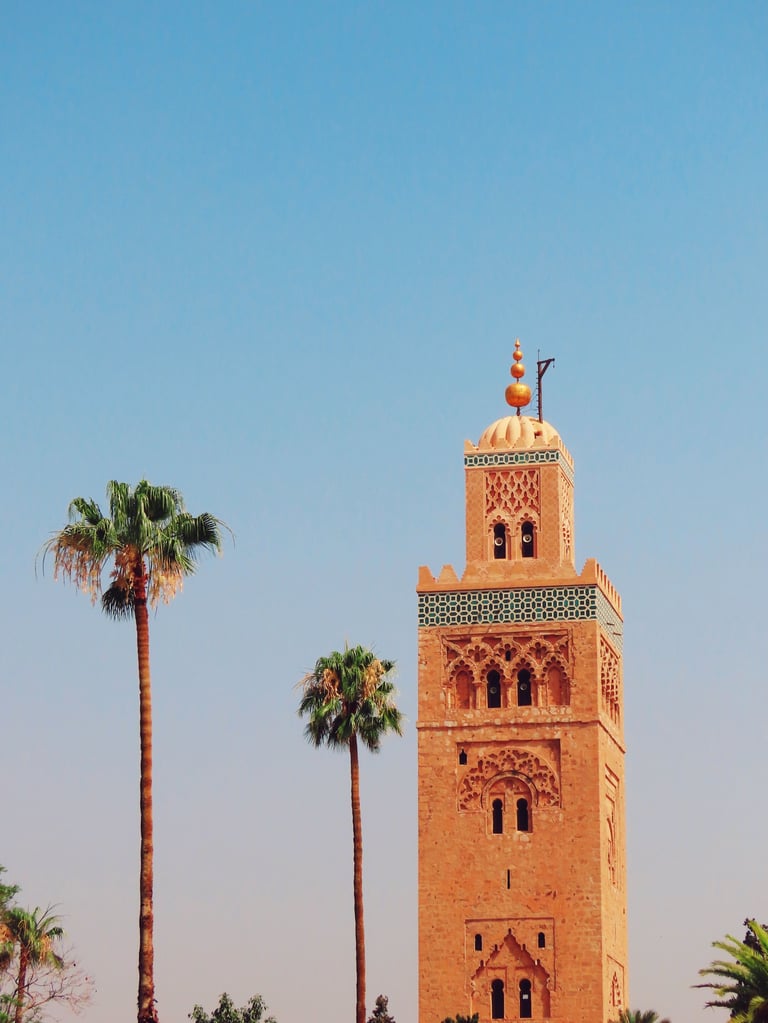 Ornate terracotta minaret tower with geometric patterns and crescent finial, surrounded by palm trees against a clear blue sky