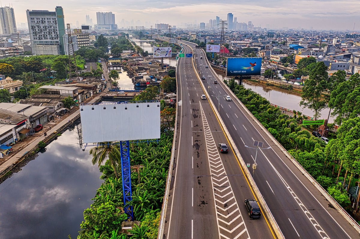 Aerial view of an elevated highway with vehicles, surrounded by urban buildings, green vegetation, and waterways.