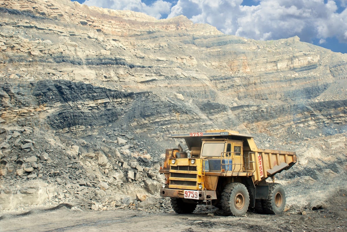 Yellow dump truck parked in front of a large quarry with exposed rock layers and layered mountain terrain