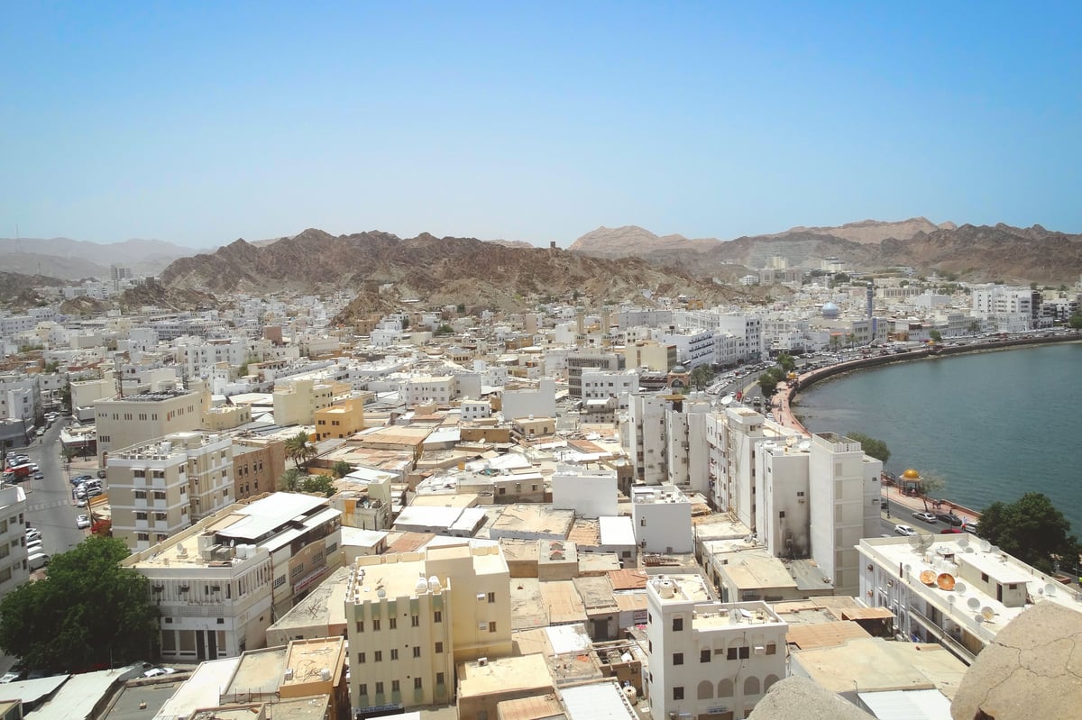 Elevated view of a Middle Eastern coastal city with tan and white buildings, waterfront, and rocky mountains under clear blue sky