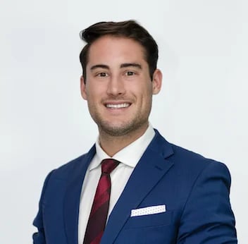 Professional headshot of a man in a navy blue blazer, white dress shirt, and burgundy tie against a light gray background