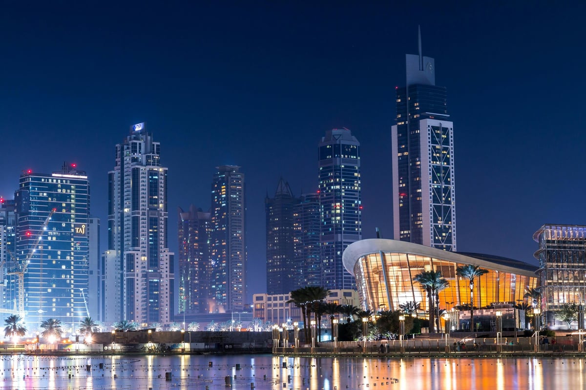 Twilight cityscape of Dubai skyline with illuminated skyscrapers reflected in waterfront, glowing concert hall in foreground
