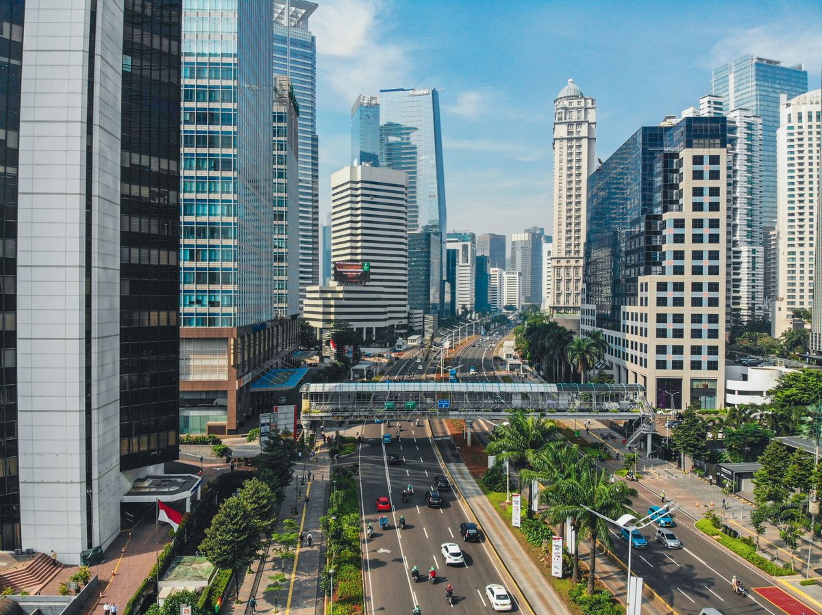 Aerial view of downtown Jakarta cityscape with modern skyscrapers, multi-lane streets, and green landscaping under clear blue sky.