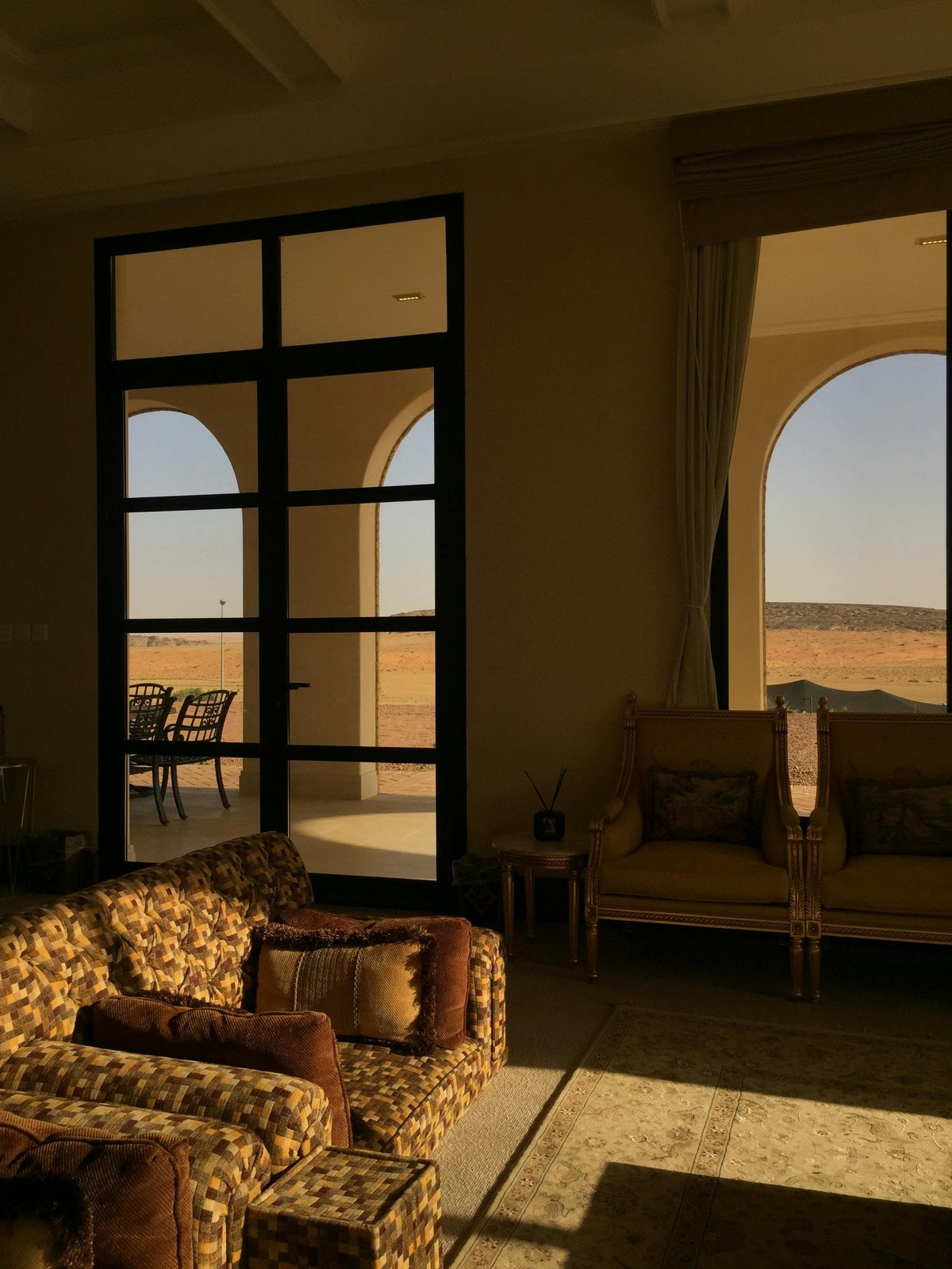 Interior room with patterned armchair and wooden furniture overlooking desert landscape through large black-framed windows and arched opening