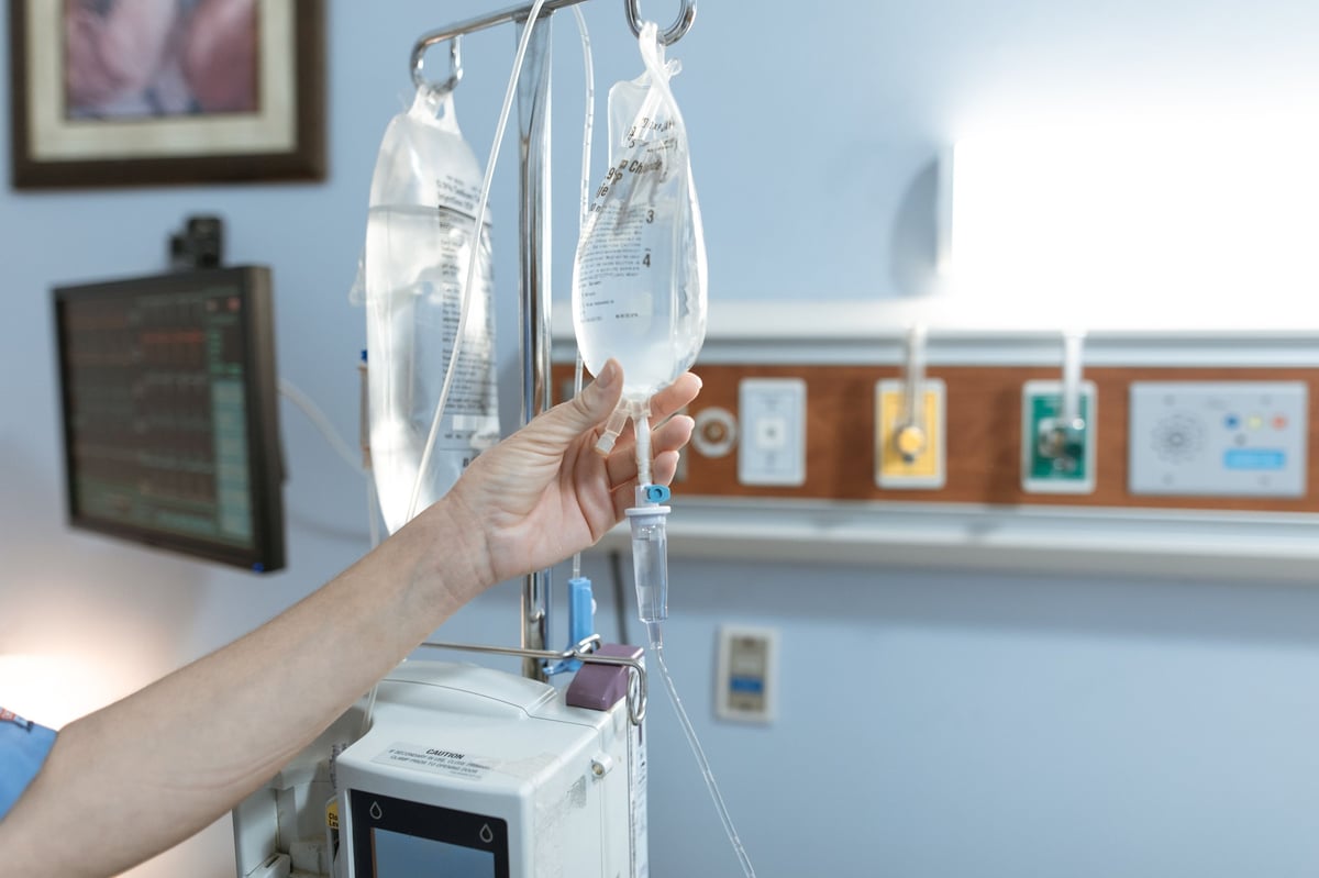 Hand adjusting an IV drip bag hanging from a medical stand in a hospital room with light blue walls and medical equipment