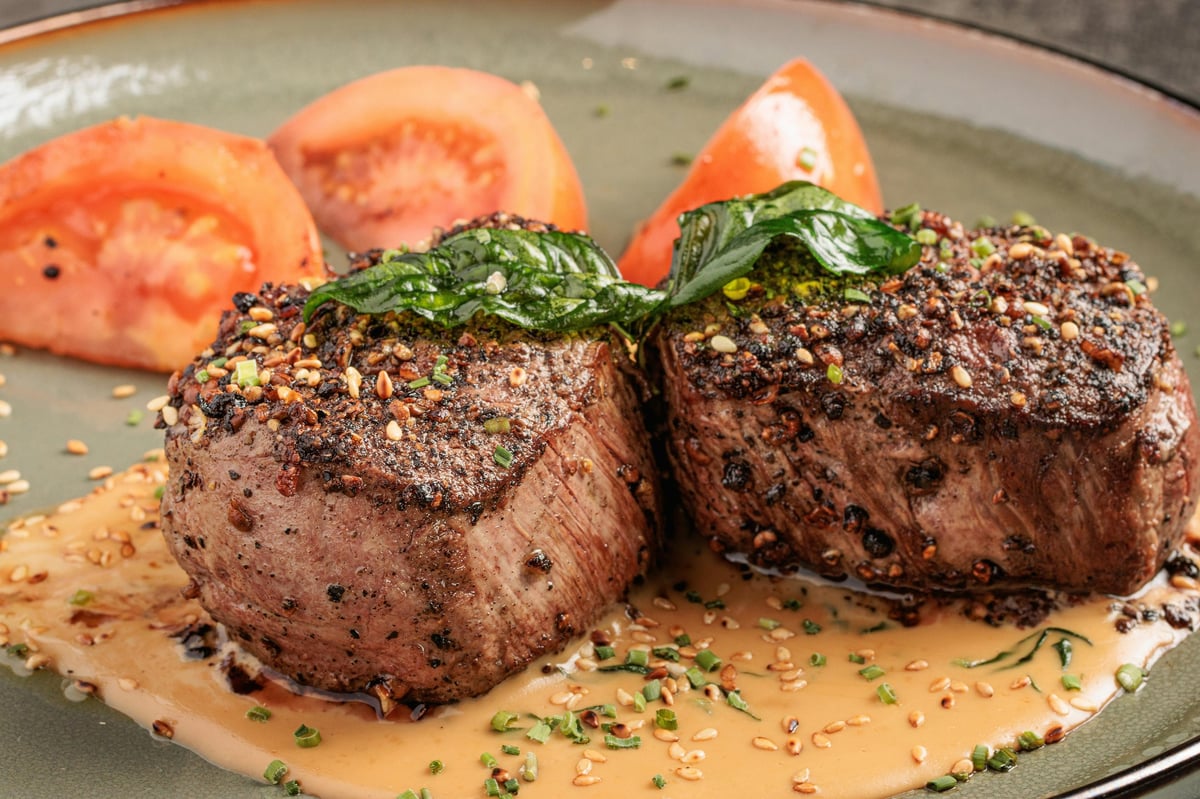 Two grilled beef steaks topped with fresh herbs on a plate with sauce and tomato slices