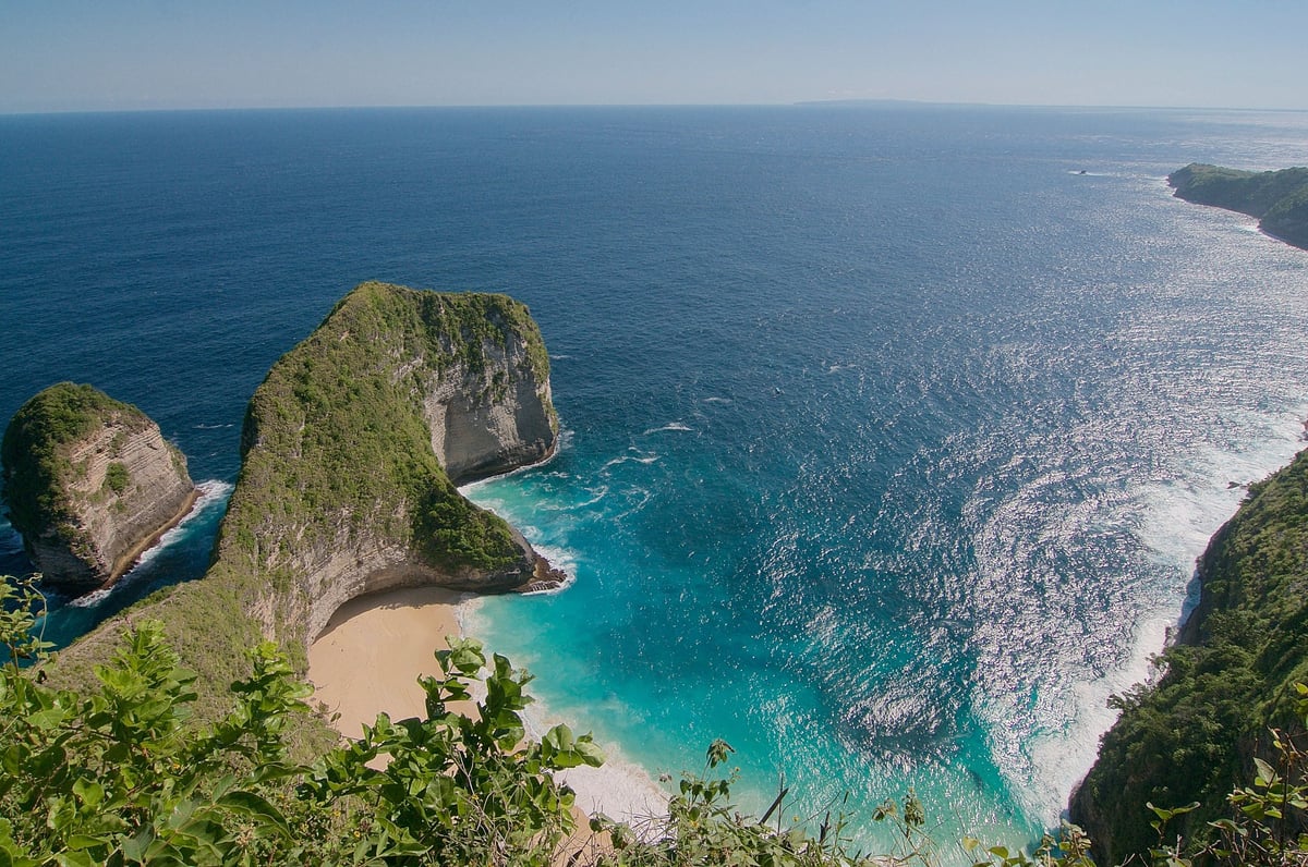 Aerial view of Kelingking Beach in Bali with turquoise water, sandy cove, and dramatic green cliffs surrounded by ocean.