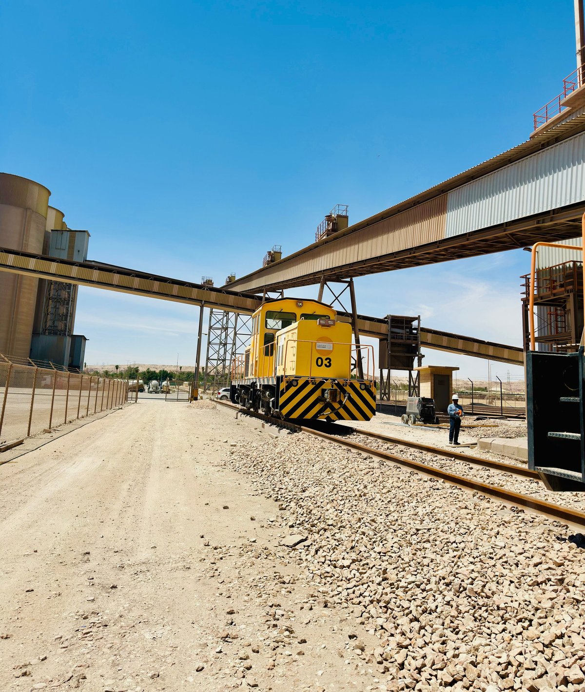 Yellow and black industrial locomotive numbered 63 on railroad tracks beneath metal overpasses at an industrial facility