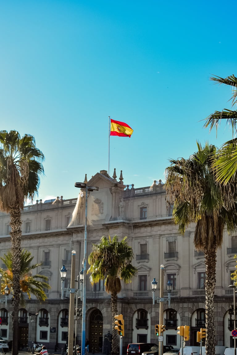 Spanish flag flying above a historic building with ornate architecture and palm trees in Barcelona