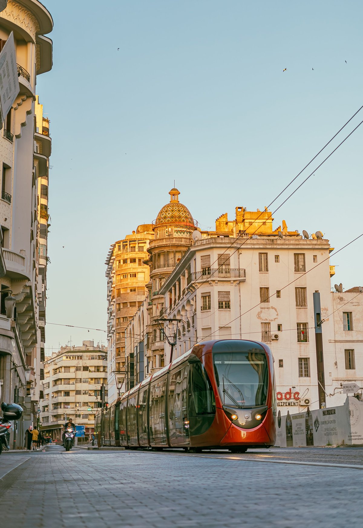 A green and red tram traveling on a street in Alexandria, Egypt, with ornate colonial buildings and a golden-domed mosque visible in the background