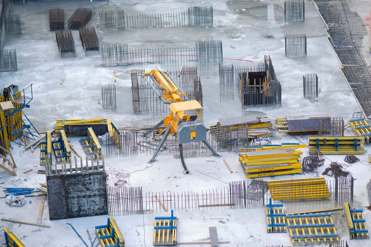 Aerial view of construction site with yellow machinery, concrete foundation, steel reinforcement, and equipment scattered on snowy ground