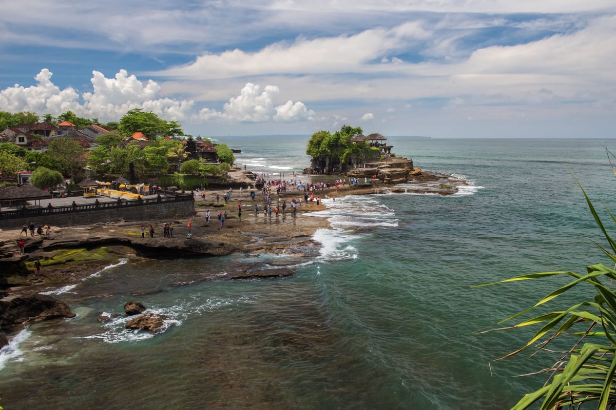 Coastal rocky cliff with a temple overlooking turquoise ocean waves under blue sky with white clouds.