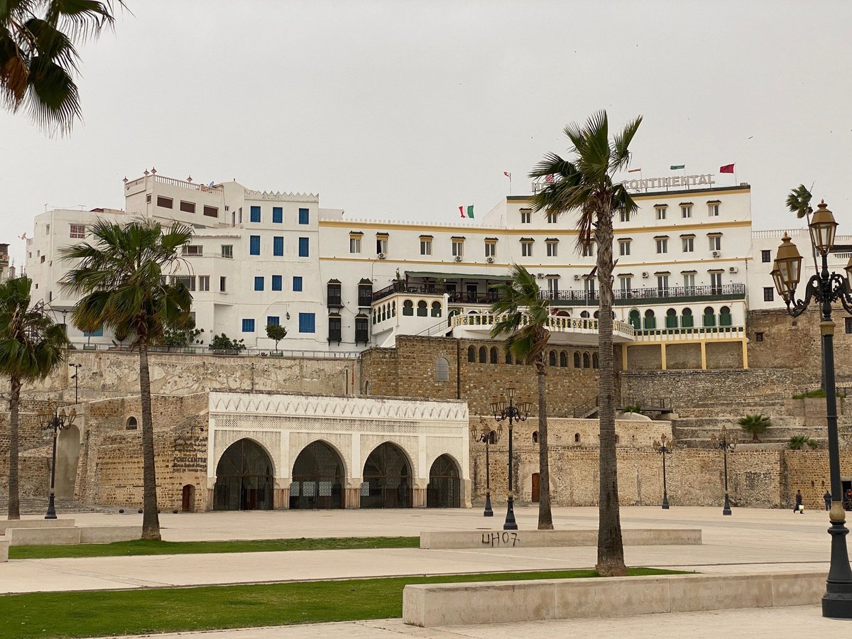 Historic stone fortress wall with arched gates and palm trees, colonial buildings visible above, green plaza in foreground