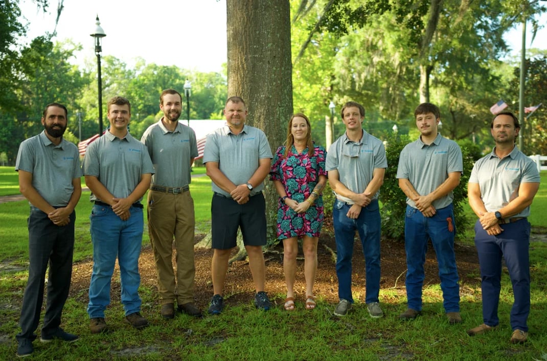 Eight people standing in a line outdoors in a park, posing in front of a large tree with green grass and sunny surroundings