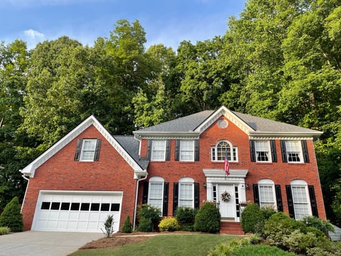 Two-story red brick colonial house with white trim, attached garage, and landscaped front yard lined with green trees