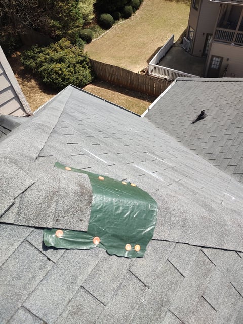 Aerial view of residential roof with gray asphalt shingles and green tarp weighted with sandbags, overlooking backyard with fence and tree