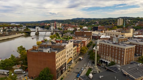 Aerial view of downtown riverfront area with brick buildings, river, and hills under partly cloudy sky