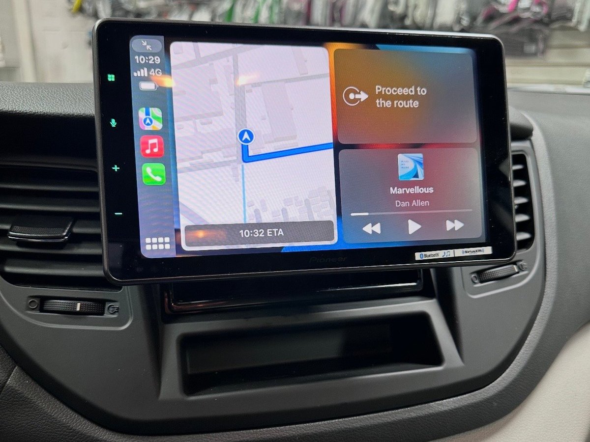Car touchscreen infotainment display showing GPS navigation map with blue route and Proceed to the route instruction, media controls visible.