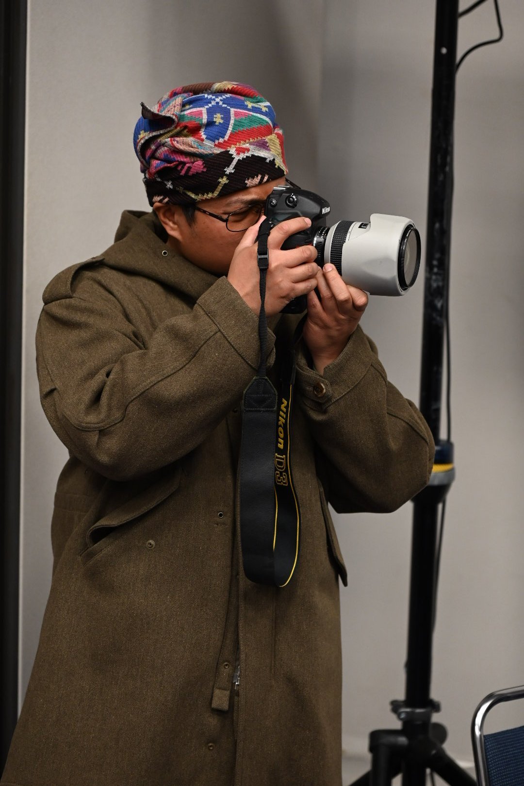 Person wearing olive jacket and colorful headwrap holding a digital camera with telephoto lens, photographing indoors