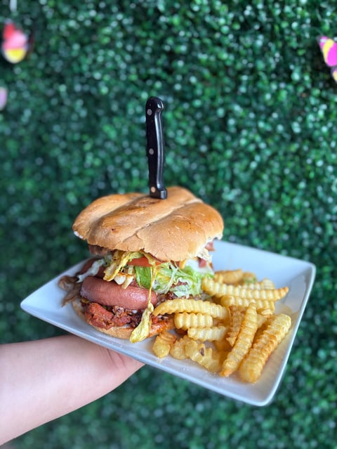 Hand holding a white plate with a gourmet burger topped with bacon and greens, accompanied by waffle fries, with a green bokeh background
