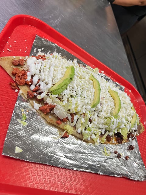 Huarache on foil and red tray topped with chorizo, shredded cheese, lettuce, avocado slices, and crema