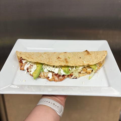 Hand holding a white rectangular plate with a folded flour tortilla quesadilla filled with chicken, melted cheese, lettuce, avocado, and crema