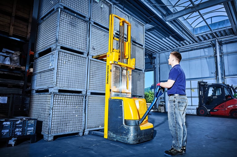 Worker operating a yellow pallet lifter in an industrial warehouse with stacked crates and forklift in background