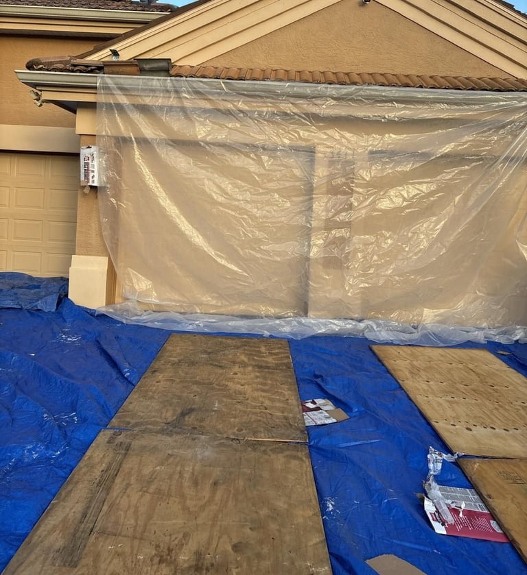 Garage wall wrapped in plastic with plywood and blue tarps protecting the driveway during a reroof