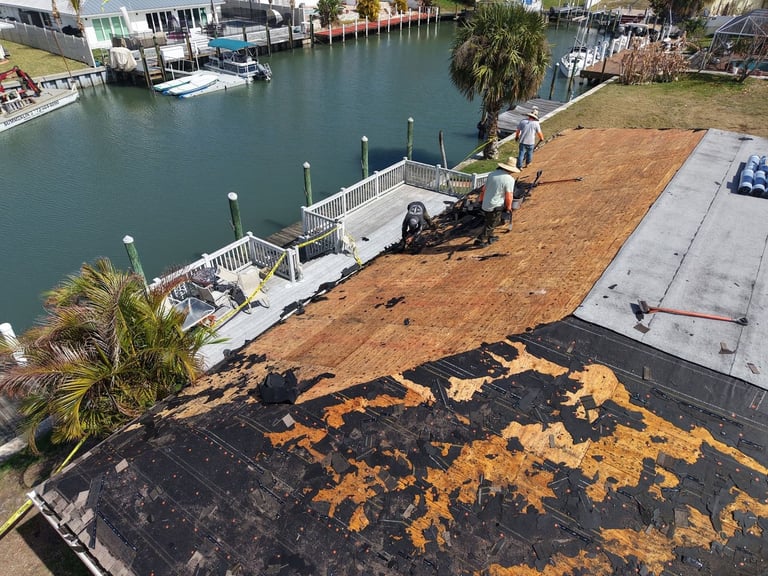 Drone view from the street of a home during shingle roof replacement, blue tarps over landscaping and a work truck in the driveway