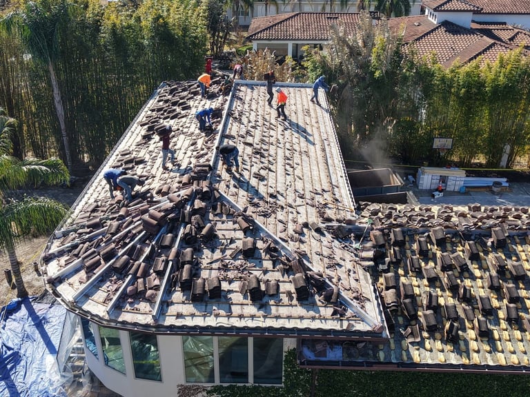 Roofing crew removing old concrete barrel tiles from a hip roof, exposing the underlayment and foam adhesive pads