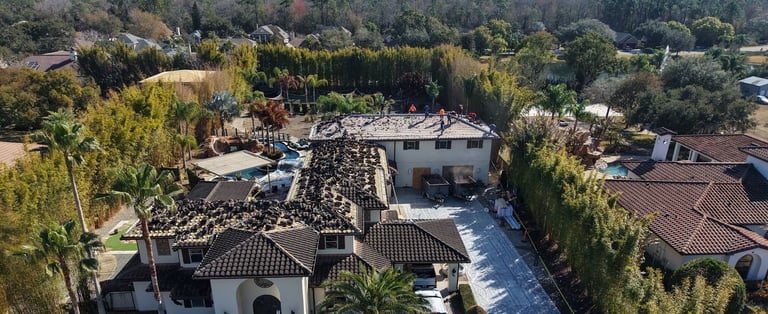 Aerial view of a large two-story home with a concrete tile roof partly torn off and roofers working on the upper roof