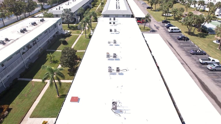 Drone view down a long white TPO flat roof with AC condensers on a two-story apartment building