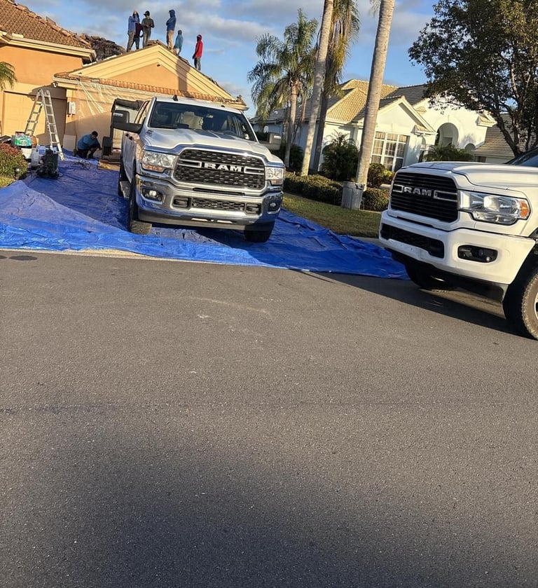 Roofing crew on a tile roof while blue tarps protect the driveway and lawn under work trucks