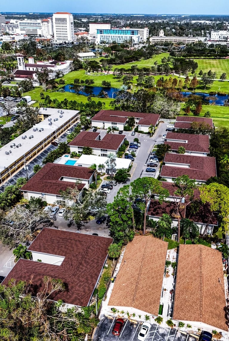 Drone view of condo buildings with red-brown architectural shingle roofs, a pool and a park beyond