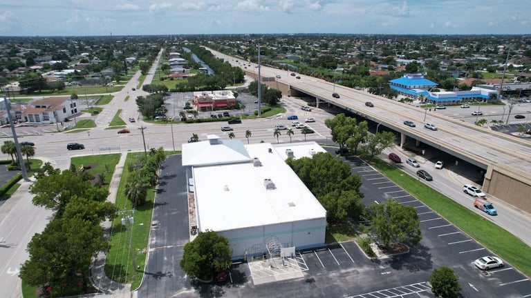Wide aerial of a commercial building with a white flat roof and silver metal hip roof next to a highway in Cape Coral, FL