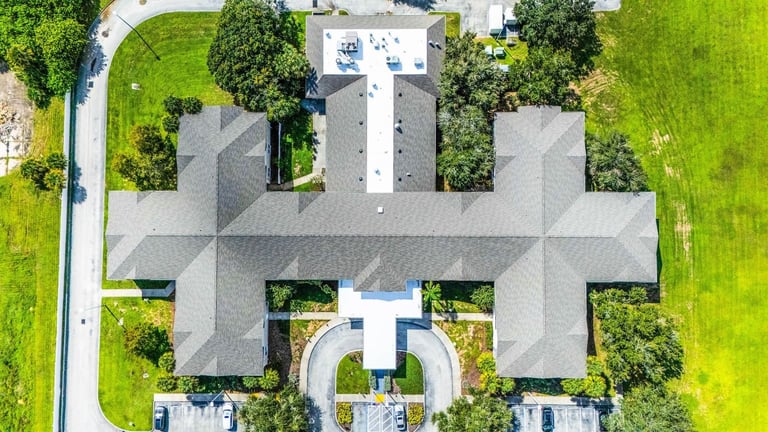 Top-down drone view of a cross-shaped care facility with a new gray shingle roof and white flat roof sections