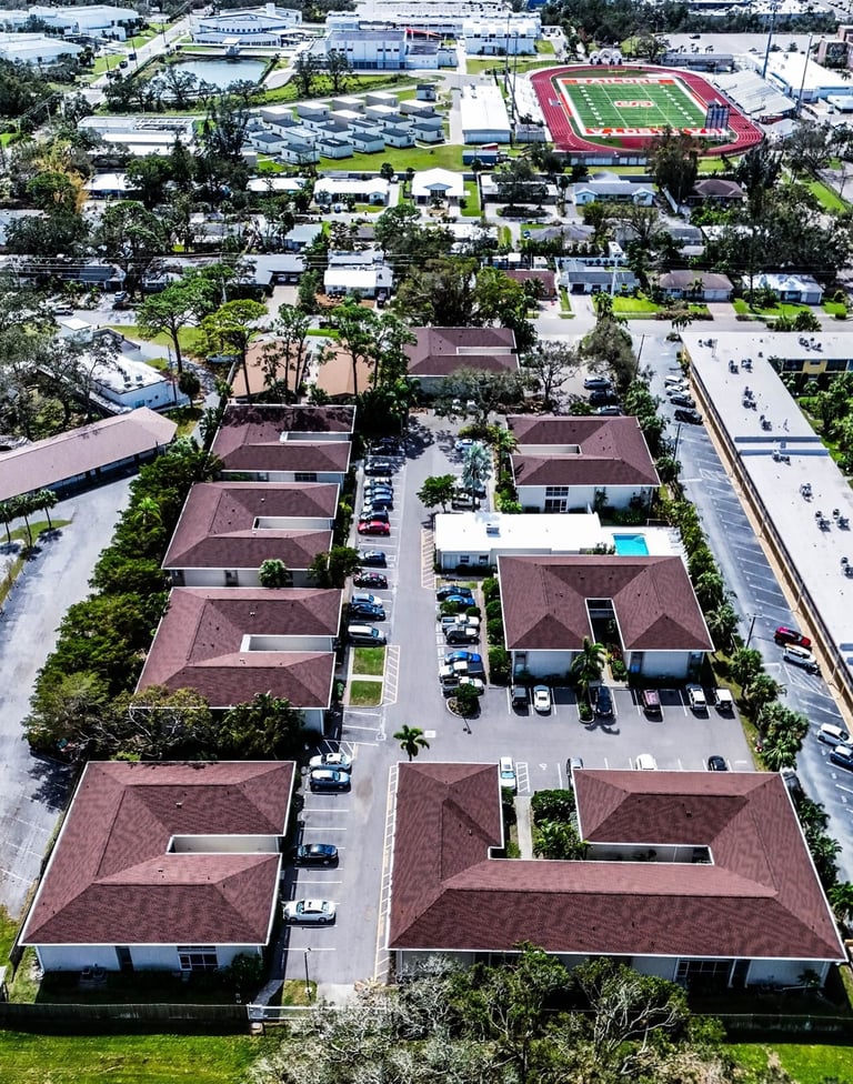 Overhead drone view of red-brown hip shingle roofs lining the parking area of a multifamily condo complex