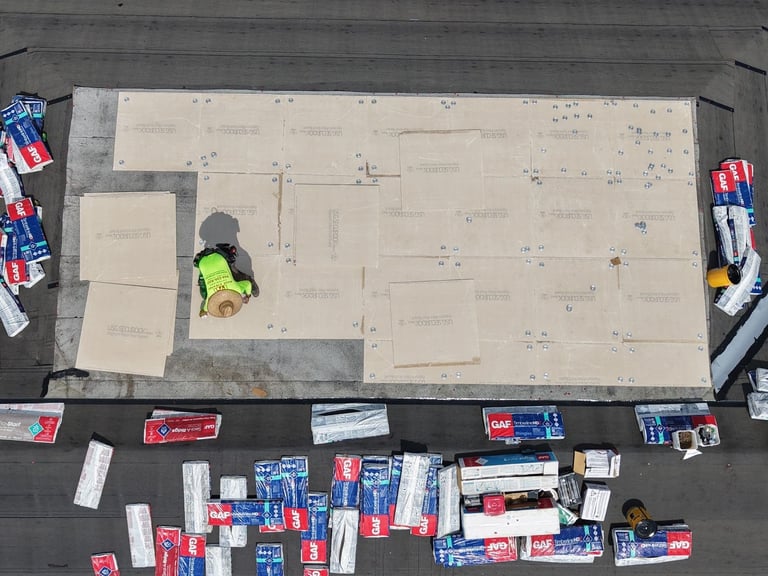 Roofer fastening cover board on a low-slope flat roof section, surrounded by underlayment and stacked shingle bundles