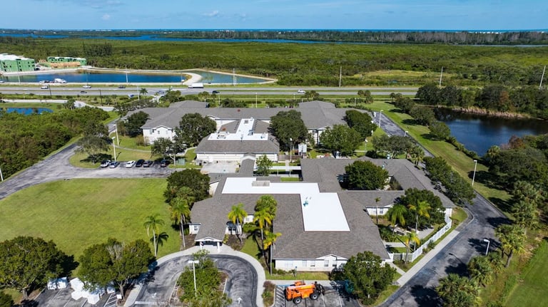 High aerial of a multi-wing care facility with new shingle roofs and a white flat roof, lakes beyond