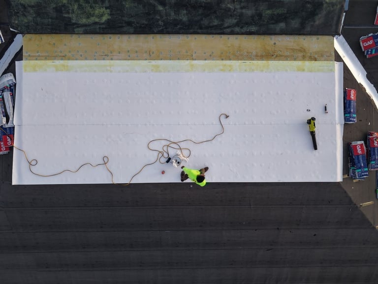 Aerial view of roofers installing a white TPO membrane on the flat roof section of a shingle roof