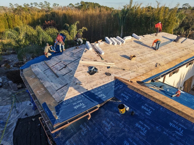Roofers rolling out peel-and-stick underlayment over the roof deck, with tile battens and supplies staged