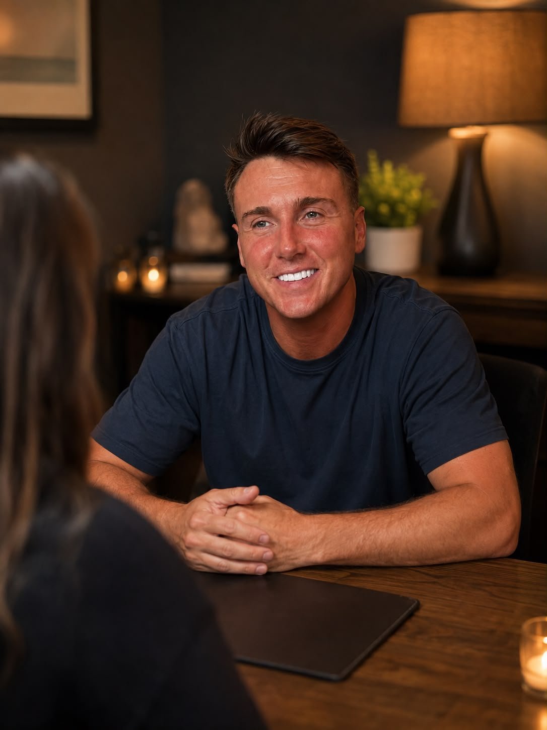 Smiling man in navy t-shirt sitting at wooden desk in warm office setting, speaking with a client during a one-to-one private reading
