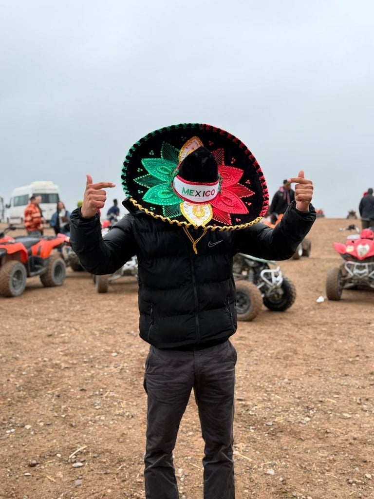 Person wearing large colorful Mexican sombrero with arms raised on desert terrain with ATVs in background