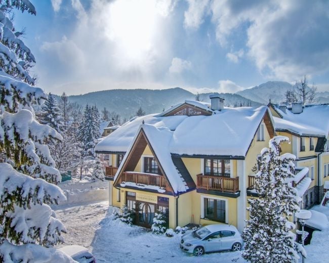 Snow-covered yellow house with brown trim in a mountain valley with pine trees and mountains in the background